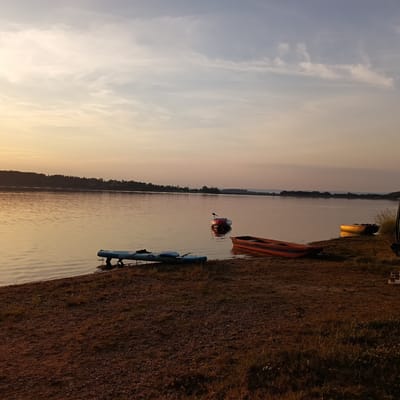 The image depicts a serene lakeside scene during sunset, with a gentle golden light illuminating the sky and water. The foreground features a rocky, grassy shoreline where two paddleboards are resting, one larger and blue, partially submerged, and another smaller one positioned further inland. A red and orange striped boat is beached on the shore in the middle ground, its hull catching the warm light. Further out on the water, a small white and red vessel, possibly a kayak or inflatable boat, floats peacefully. The water itself is calm, reflecting the soft hues of the sky. In the background, a dense line of trees forms the silhouette of the distant shore, with a hint of structures visible through the foliage. The sky is a gradient of pale yellow and soft blue, with wispy clouds suggesting a tranquil end to the day. The overall impression is one of quiet natural beauty, with the presence of recreational watercraft indicating a popular spot for outdoor activities. The provided location is the city of Jesenice, Czechia.