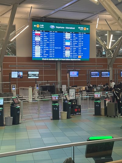 This image captures an interior view of an airport departure hall, specifically focusing on a row of check-in counters. The scene is brightly lit, suggesting it is daytime, likely mid-morning or afternoon given the flight schedule displayed on a prominent screen.

The primary subjects are the people interacting at the check-in counters and the objects associated with air travel. Several individuals, presumably travelers and airport staff, are visible. Some passengers appear to be standing in line or at the counters, while staff members are likely behind the counters, assisting them. Luggage, including suitcases of various sizes and colors, is present, indicating active travel. The setting is clearly an airport terminal, characterized by the clean, tiled floor, the rows of check-in desks, and the large departure board.

The activity taking place is airport check-in. The departure board displays a list of international flights with their scheduled times, destinations, airlines, flight numbers, and gate information. Some flights are marked as "Delayed" or "Cancelled," adding a layer of realism to the airport environment. Signage for "AIR CANADA" and instructions like "Step 1" and "Bag Drop" further clarify the function of the area. The text on the departure board and the signage provides crucial details about the flights and airport services.

Notable details include the color-coded tiling on the floor, which creates a somewhat cheerful atmosphere. The green illuminated signs above some of the counters suggest they are operational. The overall impression is one of a busy, functional airport environment, with travelers moving through the necessary procedures before their flights. The location is identified as Richmond, Canada.