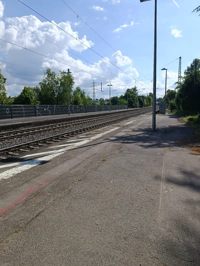The image depicts a train platform in Eltville, Germany. The scene is taken from the platform level, facing down the tracks, with overhead power lines and supporting poles visible above. To the left of the tracks, a dark wooden fence runs alongside, adorned with graffiti. Two sets of train tracks stretch into the distance. The platform itself is paved with a rough gray asphalt, marked with faded white and red lines near the edge. A tall, cylindrical metal pole stands on the platform, casting a shadow. To the right, vegetation and trees frame the view, suggesting the edge of the station area. The sky above is bright blue with puffy white clouds.