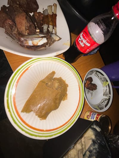 The scene depicts a tabletop arrangement of food and drink in Debasco, Nigeria. In the upper left, a white, irregularly shaped serving dish contains a substantial quantity of dark, cooked meat, including ribs with visible bones. Below this, a white bowl with green and orange concentric stripes on its rim holds a large, light brown, paste-like food item, consistent with a traditional Nigerian meal component. To the right, a smaller white bowl with a floral/geometric pattern contains a few dark, small pieces of cooked food. A plastic bottle of "American Cola" with a red cap is partially visible, positioned diagonally on the table, indicating it has been consumed. Further right, a yellow can labeled "Three Crowns" is present, alongside a dark container holding a pale, textured substance. The table surface is light brown wood, and the background is dark, suggesting an indoor setting with ambient lighting. No individuals are present in the image.
