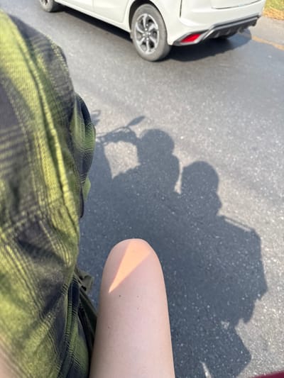 A high-angle shot shows a person wearing a green and black plaid shirt with part of a leg visible. The person's shadow, along with the shadow of a bicycle, is cast on the asphalt pavement. A white car is partially visible on the left side of the frame. The car's back tire is clearly seen.