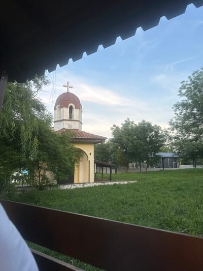The image captures an outdoor scene in Bliznatsi, Bulgaria, framed from a low perspective. The top portion of the image is dominated by a dark, serrated overhang, consistent with a roof or awning structure. A dark wooden railing or bench back extends across the lower foreground, partially obscuring the bottom left corner, where a segment of white fabric, likely clothing, is visible.
The central subject is a small, light-colored chapel or church, positioned towards the left-center of the frame. This structure features a prominent reddish-brown dome-shaped roof (cupola) surmounted by a white cross. Below the dome is a white bell tower with arched openings. The main body of the building has yellowish walls and a terracotta-tiled awning extending over a covered entrance area. Dense green foliage from various trees and bushes partially envelops the chapel, particularly on its left side, where long, drooping leaves are visible.
To the right of the chapel, a wide expanse of green grass covers the midground. Several deciduous trees with full, green canopies are scattered across this grassy area. In the background, beyond the trees, a modern-looking structure with a dark, possibly metallic, roof and dark walls is visible. A low wall or fence line appears to demarcate sections of the landscape in the distance.
The sky in the background is clear blue with some faint, wispy clouds, suggesting either late afternoon or early evening light. No individuals are clearly visible in the scene, although the white fabric in the lower left implies the presence of an observer or person near the camera.