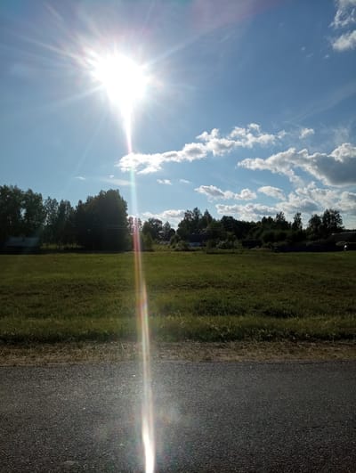 This media file depicts a scenic outdoor scene, likely in a rural setting, with a bright, sunny day. The dominant elements are a vast blue sky dotted with fluffy white clouds, a vibrant green grassy field, and a dark asphalt road running along the edge of the field. The sun is prominently visible, casting a strong glare and lens flare across the image, indicating it is likely mid-day.
In the background, a line of lush green trees forms a natural border between the field and the sky. There are hints of structures within or behind the trees, possibly buildings or outbuildings, suggesting human presence or habitation in the vicinity. The overall impression is one of natural beauty and tranquility, with the bright sunlight contributing to a cheerful and energetic atmosphere.
The image is oriented vertically, as if taken while standing and looking upwards or from a slightly elevated position. The lighting suggests it is a clear day with good visibility. There is no visible text in the image. Given the location context of Гарбавіцкі сельскі Савет, Belarus, the scene is likely representative of the Belarusian countryside.