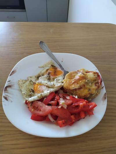 An overhead shot displays a white plate filled with a meal placed on a wooden table. The plate is decorated with a brown floral pattern along its edge. On the left side of the plate are two fried eggs, seasoned with black pepper. In the center, a portion of chopped red bell peppers and tomatoes, mixed with diced onions and herbs, forms a salad. To the right, a round, layered dish is topped with melted golden-brown cheese and appears to be a baked item, possibly a meatloaf or casserole with tomato slices. A silver fork rests on the plate, its tines pointing towards the top right corner of the frame. The background shows a portion of a kitchen counter with cabinets. This image was taken in Bischofsheim, Germany.