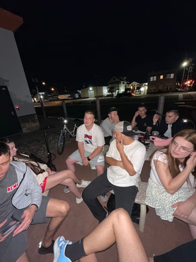 In Ocean City, United States of America, a group of seven young adults, appearing to be in their late teens or early twenties, are gathered outdoors at night. The setting appears to be a patio area, possibly associated with a commercial establishment, with a building facade visible on the left. The ground is paved with what looks like reddish-brown brick or tile, and gravel is visible in the background.
The group is casually dressed. Several individuals are seated, with two women and five men. One man in the foreground, on the left, is wearing a grey jacket with "ACMI" branding and grey shorts. Next to him, a woman is seated with her legs crossed, wearing a tan hoodie and red shorts. Another woman, on the far right, is wearing glasses and a floral skirt. The men are dressed in t-shirts and shorts, with one wearing a baseball cap.
Objects in the scene include a bicycle with a blue rim, an electric scooter, and a table around which some individuals are seated. There are also drinks on the table. The lighting suggests it is nighttime, with artificial lights illuminating the area and the background showing some distant buildings and streetlights. The overall atmosphere appears relaxed and social.