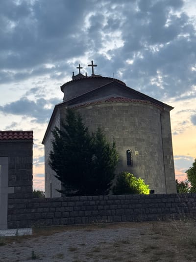 An imposing, round, stone-built church with a tiled roof stands prominently against a dramatic sky filled with dark, swirling clouds. Three crosses adorn the top of the structure, with two clearly visible at the apex. A small, arched window with dark bars is set into the stone facade. To the left, a smaller, rectangular building with a similar tiled roof and a stone wall with a cross emblem is visible. A low, dark stone wall runs across the foreground, partially obscuring the view of a sparse, dry grassy area. A tall, dark green conifer tree stands directly in front of the main church building. To the right of the church, a patch of lush green foliage is brightly illuminated by a light source, creating a stark contrast with the surrounding darker elements. The background shows a sliver of a warm, colorful sunset sky peeking through the clouds. The scene appears to be set in an outdoor, perhaps rural, location, likely in the city of Zagora, Montenegro, given the architectural style and the overall atmosphere. There are no people or active events depicted in the image.