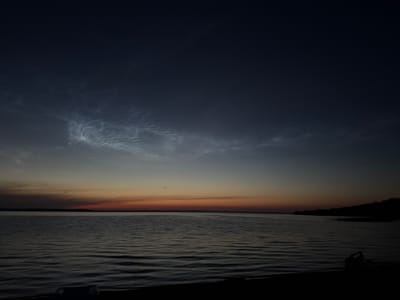 The image shows a serene twilight scene over a large body of water, likely a lake or river, with a dark, silhouetted shoreline in the foreground. The sky transitions from a deep indigo at the top to shades of orange and red near the horizon, indicating either sunset or sunrise. Wispy, luminous clouds, possibly noctilucent clouds, are visible in the upper portion of the sky, catching the fading light. The water is calm with gentle ripples reflecting the colors of the sky. On the far bank, a dark, treed hill is visible. In the immediate foreground on the right side of the frame, two indistinct human figures are visible in silhouette, one appearing to be sitting or crouching near the water's edge. The overall impression is one of tranquility and natural beauty. The location is identified as Володарский ауылдық округі, Kazakhstan.