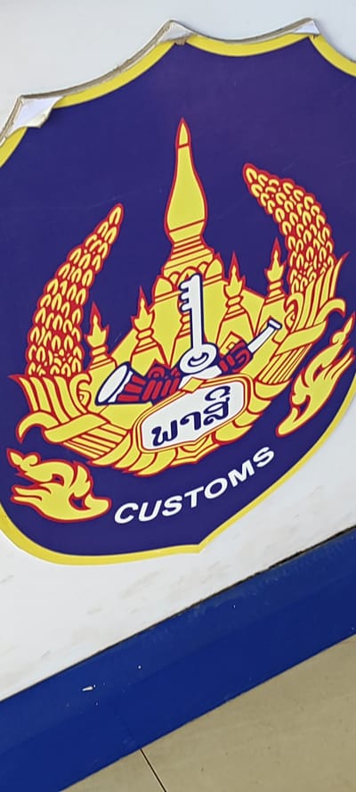 This image shows a close-up of a logo on a surface. The logo is shield-shaped with a purple background and a yellow border. It features a central design in yellow and red depicting a temple-like structure, a key, and what appears to be ears of rice or wheat. Below the central design, there are two lines of text. The top line is in a script that appears to be Lao, and the bottom line reads "CUSTOMS" in white English letters. The logo shows some wear and tear, with peeling edges. The background outside the logo is a white surface with a blue strip along the bottom edge. The image is slightly angled and vertically oriented. There are no people or discernible environmental details indicating a specific location within the city of Xayden, Laos. The image focuses solely on the logo and the surface it is affixed to.