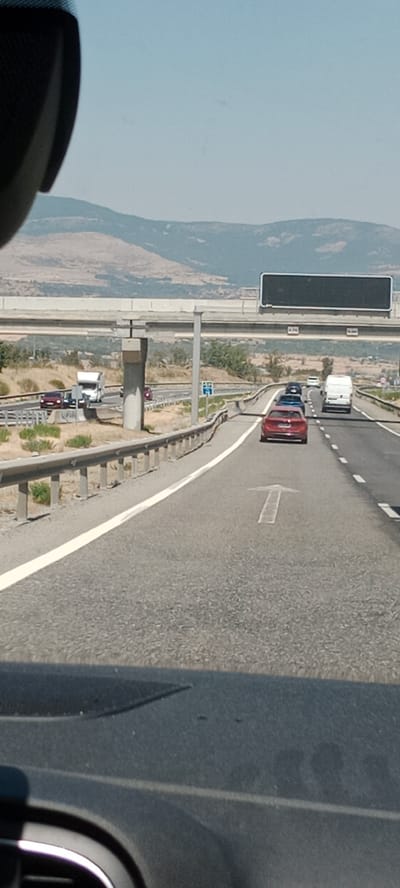 The image depicts a view from inside a vehicle traveling on a highway near Buitrago del Lozoya, Spain. The highway has multiple lanes, with a concrete barrier and guardrail separating the lanes from the shoulder. Several vehicles are visible on the highway, including cars, a truck, and a white van. The landscape in the background consists of dry, rolling hills and mountains under a clear blue sky. A pedestrian overpass is visible in the midground, with a large black digital sign above it. The road surface is asphalt with white lane markings, and a white arrow pointing forward is painted on the right lane. The interior of the vehicle, including the dashboard, is partially visible in the foreground.