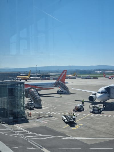 The image shows an aerial view of the EuroAirport Basel-Mulhouse-Freiburg, specifically the part located in Saint-Louis, France. Visible in the foreground are aircraft on the tarmac. An EasyJet plane with orange tail and lettering is prominent. A yellow DHL aircraft is also visible, docked at a terminal. A Lufthansa aircraft is also visible docked at a terminal. There are various ground service vehicles parked around the planes. The tarmac is marked with painted lines and numbers. In the background are mountains and a blue sky with wispy clouds. The image appears to be taken from inside an airport terminal, looking out through a window, as a part of the window can be seen reflected.