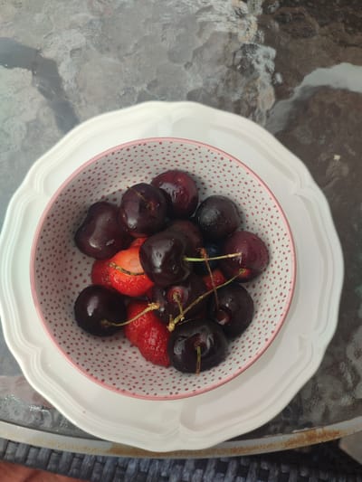 A top-down shot shows a small, patterned bowl filled with cherries and strawberries. The bowl is resting on a larger, white, scalloped plate, which is on a textured, dark surface. The background is a blurry, reflective surface, possibly a glass table. The fruits are dark red and glossy, with some water droplets visible on their surfaces. The strawberries are smaller and brighter red, with some green stems still attached. The overall lighting suggests it might be daytime, possibly outdoors given the reflective surface, but the exact weather conditions are not discernible. The scene is simple and focused on the fruit. No people, text, or specific time of day indicators are visible. The location context provided is Longueuil, Canada, which is not directly evident from the image itself but adds context to the setting.