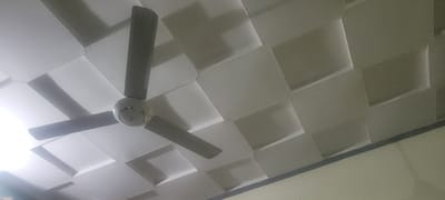 This media file shows a low-angle view of a white, textured ceiling with a modern, geometric design. The ceiling is made up of recessed square panels that create a dynamic, faceted surface with shadows highlighting the depth. A ceiling fan with three gray blades and a white central motor housing is prominently featured, positioned in the upper left quadrant of the image. One blade is angled towards the viewer, and the other two extend out of frame. The lighting suggests it is daytime, with a bright light source, possibly natural light from a window or an overhead lamp, illuminating the scene from the left and casting soft shadows on the ceiling. The overall impression is one of a clean, contemporary interior space. The location context provided is Kipala Mpakani, Tanzania. There is no visible text in the image.