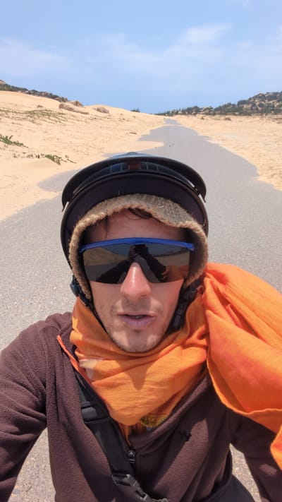A selfie taken in Phuoc Dinh, Vietnam, shows a person wearing a brown fleece jacket, an orange scarf, a black helmet, and blue-framed sunglasses. The background consists of a sandy area, possibly dunes, meeting a clear blue sky. The person is likely standing on a paved surface.