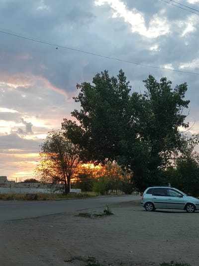 The image depicts a roadside scene at dusk in Kurchatov, Kazakhstan. A light blue station wagon is parked on a dusty, unpaved shoulder next to an asphalt road. Large trees with dark green foliage dominate the left side of the frame, with their branches partially obscuring the sky. The sky itself is filled with dramatic clouds, with streaks of orange and pink from the setting sun visible through the trees. The overall mood is serene and perhaps a bit melancholic, indicative of the end of the day. Power lines are visible stretching across the sky. In the distance, there are hints of low-lying structures, possibly a fence or the remnants of buildings. The ground is dry and textured, with scattered weeds and debris. The time of day is clearly sunset, given the colors in the sky and the soft, fading light.