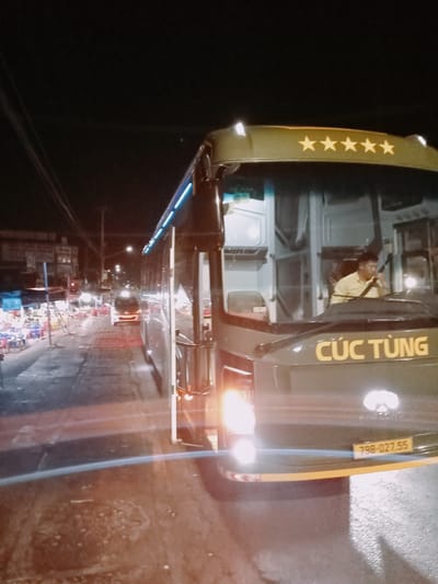 A nighttime shot depicts a Cúc Tùng bus, its headlights on, positioned on a street in Ea Drăng, Vietnam. The bus, dark green with the name "Cúc Tùng" in yellow on its front, also features five stars above the windshield. The license plate reads "79B-027.55". A man in a light-colored shirt is seen driving the bus. The street is wet and reflects light. On the left, tables and chairs line the sidewalk, suggesting a restaurant or gathering place. In the distance, a few vehicles and streetlights illuminate the street.