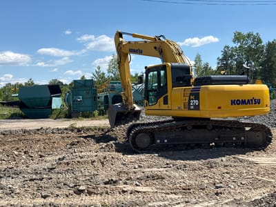 This media file displays a Komatsu PC 270 LC excavator parked on a gravelly, uneven terrain, likely a construction or demolition site. The excavator is yellow with black accents, featuring the "KOMATSU" logo prominently on its boom and body. The machine's tracks are visible, as is its bucket, which is resting on the ground.
In the background, several large, industrial-looking green machines are present, possibly part of a processing or sorting facility. The setting suggests an outdoor industrial area. The sky is blue with scattered white clouds, indicating a bright, clear day, likely daytime given the strong shadows.
No people are visible in the media file. The overall mood is one of industrial activity or preparedness, with the heavy machinery suggesting ongoing or imminent work. The location context is Makariv, Ukraine, though this cannot be visually confirmed from the image alone.
Visible text includes "KOMATSU" on the excavator's boom and body, and "PC 270 LC" on the side of the excavator's chassis. There are also smaller warning or identification labels on the machine.