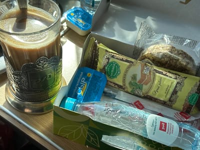 The image shows a close-up of a meal and drink setup, likely on a train or similar travel setting. A glass of coffee or milk is in a silver-colored holder. Next to the drink is an opened cardboard box containing several items: a small container of processed cheese, a bottle of water with a label indicating it was made for a "Federal Passenger Company" and an item labeled "Belyovskaya Pastila" which is apparently made from baked Antonov apples. There is also some crispbread. The objects are situated on a wood-like surface. The location in the city of Zainsk, Russia, is not discernible from the image content.