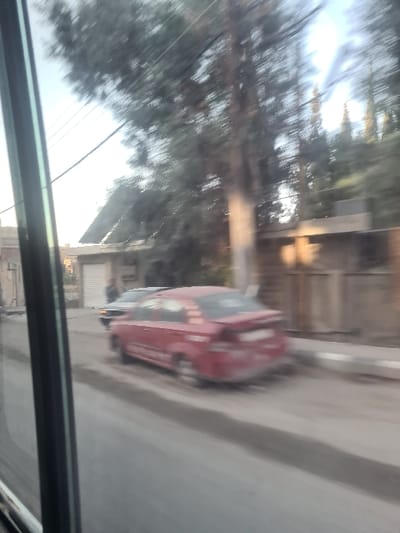 A blurry, angled shot taken from inside a vehicle. A red car is parked on the side of a street with a building next to it, and trees behind it. The image is heavily blurred, potentially due to movement. The frame of the window through which the picture is being taken is visible. The outdoor lighting appears to be daytime or dusk.