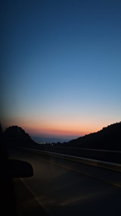 The image captures a scenic view from a vehicle traveling along a road at dusk. The sky displays a gradient of colors, transitioning from a deep blue at the top to hues of orange and pink near the horizon, indicative of a sunset or twilight. Silhouetted against the colorful sky are the dark outlines of rolling hills or mountains on either side of the road. The road itself is paved and appears to be a highway or a major route, with a guardrail visible along the side. The foreground is dominated by the dark interior of the vehicle, with a portion of the side-view mirror visible on the left. In the distance, lights from a settlement or town can be faintly seen nestled between the hills. The scene is devoid of any people or specific actions, presenting a serene and tranquil landscape at the end of the day. The location is identified as Malo Buchino, Bulgaria.