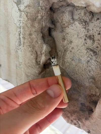 A hand holds a cigarette, its glowing tip touching a rough, textured surface. The cigarette appears to be partially lit, with ash visible at the end. The hand is a light skin tone, with a manicured fingernail. The cigarette has a gold-colored filter and a white band. The background is a mottled gray and beige, with a rough, uneven texture that suggests stone or concrete. There are crevices and dark spots within the surface, suggesting age or weathering. The lighting is bright, casting subtle shadows. The overall scene is close-up, focusing on the interaction between the cigarette and the textured surface. The image is taken from a slightly elevated angle, looking down at the hand and cigarette. No text is visible. The location context of Orhei, Moldova, provides a geographical setting for the image.
