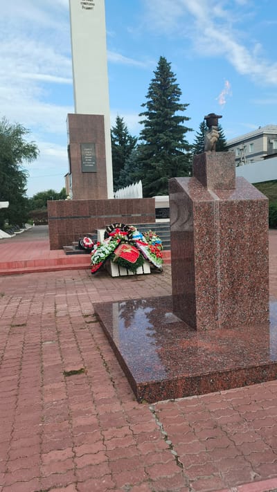 This image depicts a war memorial located in Buguruslan, Russia. In the foreground, a large granite monument with a flame on top stands prominently. To the left of this monument, a collection of funeral wreaths, some adorned with red stars, are placed at the base of a larger, white obelisk that is inscribed with text. The ground is paved with red interlocking bricks, and a few patches of grass are visible between them. In the background, evergreen trees and a modern building can be seen under a cloudy blue sky. The scene appears solemn and commemorative.