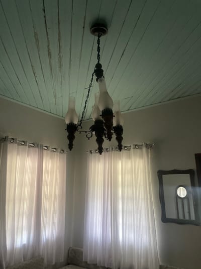 The image depicts the interior of a room located in Pirenópolis, Brazil. The ceiling consists of light blue-green painted wooden planks, exhibiting visible grain and minor surface imperfections. A dark-colored, five-arm chandelier hangs centrally, suspended by a chain. Each arm incorporates decorative dark wooden or stained elements and holds an inverted, frosted glass shade. Below the chandelier, two large windows are covered by sheer, floor-length white curtains featuring subtle vertical stripes, hung via metal grommets on a rod. Diffuse daylight illuminates the room through these curtains. On the right wall, a rectangular mirror with an ornate, dark wooden frame reflects a circular mirror and a portion of a wooden object. The lower foreground shows a section of an upholstered sofa or seating element with a light-colored, patterned fabric. No individuals or specific actions are present within the scene.