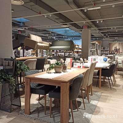 The image depicts the interior of a furniture store. In the foreground, a large wooden dining table is set with a runner, a vase with flowers, and a potted plant. Several upholstered dining chairs surround the table, including dark grey chairs with diamond-patterned seats and light beige chairs. To the left of the table, a black metal stand holds potted plants and smaller pots. In the background, various sofas and other furniture pieces are displayed, with shelving units visible further back. The store appears to have a concrete ceiling with exposed ductwork and lighting fixtures. The overall lighting suggests an indoor commercial environment. The timestamp in the lower right corner indicates the image was captured on July 24, 2026, at 10:01. There are no people visible in the image. The location is identified as Modugno, Italy.