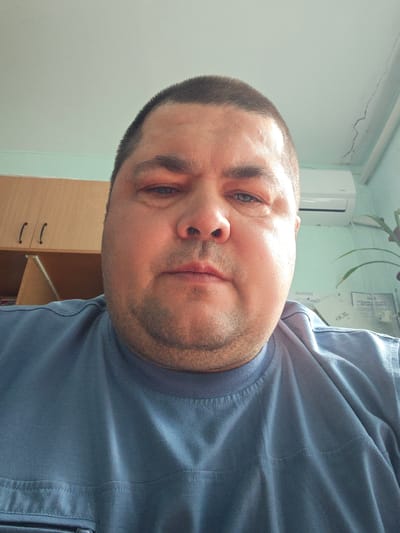 A close-up, low-angle selfie of a middle-aged Caucasian man with a buzz cut and a full face is captured. He is wearing a blue t-shirt, and the shot focuses on his face and upper chest. The background appears to be an indoor setting, possibly an office, with light blue walls and a white air conditioning unit visible on the wall. To the left, a wooden cabinet with two handles is present. The overall lighting is bright, suggesting it might be daytime. The man's expression is neutral. This image is likely taken in Sovietskyi, Ukraine, given the provided context, though no specific landmarks are identifiable.