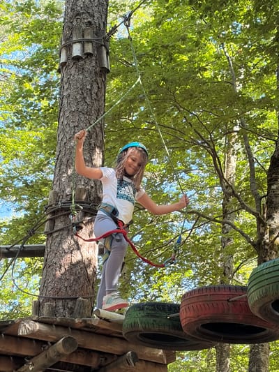 A young girl, estimated to be between 8 and 12 years old, actively participates in an aerial adventure course within a forest located in Mali Bostur, Montenegro. She is positioned on a wooden platform attached to a large tree trunk, which has dark, textured bark. The girl wears a blue protective helmet, a white short-sleeved t-shirt with a teal and black Adidas logo on the chest, light grey leggings, and light-colored athletic shoes with pink accents. A red and blue safety harness is visible around her waist and legs. She grasps a thick green safety rope with both hands, extending her arms upwards. Her facial expression indicates a smile. The tree trunk is outfitted with horizontal wooden logs secured by ropes, forming part of the course infrastructure. To the right of the girl, a series of recycled tires, painted red, green, and black, are suspended by cables, forming a subsequent obstacle. The background is dominated by lush green foliage of numerous trees, indicating a dense forest environment, with intermittent patches of blue sky visible through the canopy. Sunlight illuminates parts of the scene, creating bright highlights on the leaves and the girl's clothing.
