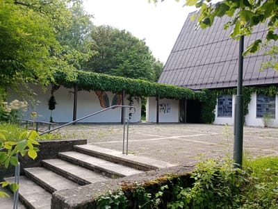 The image depicts an outdoor area with a modern building featuring a large, grey, angular roof. A white wall runs along the side of the building, partially covered in dense green ivy, with a mural of large, colorful wings visible on its surface. A metal railing with steps leads up to this area. The ground is paved with concrete squares, with some weeds growing in the cracks. Trees and lush green foliage surround the building. The scene appears to be the exterior of a public building, possibly a church or community center, in Brannenburg, Germany. There are no people visible in the image.