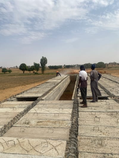 The image displays an active construction site in Wuciciri, Nigeria. A long, partially completed concrete structure, likely a culvert or bridge deck, extends from the foreground towards the mid-distance. This structure is composed of numerous light-grey precast concrete slabs, many with exposed rebar loops. A central, longitudinal channel within the structure contains standing water further back.
Three individuals are present. In the mid-distance, positioned on the structure's left side, one person wearing an orange safety vest over dark clothing operates a power tool on the concrete, producing a visible cloud of white dust. To the right, two men observe this activity. The closer man wears a white and dark two-tone t-shirt, dark pants, and brown shoes, holding a dark object in his left hand. The second man, standing slightly behind and to his right, is dressed in a grey traditional tunic and a dark head covering, with his hands placed on his hips.
The background consists of dry, open land with sparse vegetation and scattered trees under a pale blue sky with wispy white clouds. The horizon appears hazy.