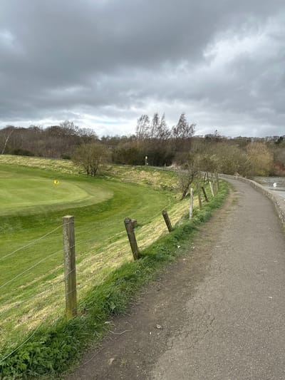 The image depicts a golf course on an overcast day. A path runs along the right side of the frame, separated from the fairway by a wooden post and wire fence. The fairway is green and well-manicured, with a yellow flag indicating the location of the hole. Trees line the left side of the frame. The sky is filled with gray clouds. The image was taken in Dunfermline, United Kingdom.