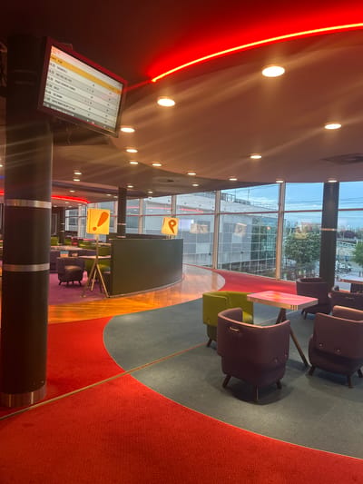 The image captures the interior of a cinema in Ivry-sur-Seine, France. Seating areas with purple chairs and colored tables are arranged on a multi-colored carpet: red, blue, and beige. A large screen displays movie showtimes, including "COCORICO 2," "THEY WILL KILL YOU," "PROJET DERNIERE CHANCE," "DOLLY," "THE DRAMA," and "SUPER MARIO GALAXY LE FILM." Several lights illuminate the space: overhead spotlights and a red neon strip along the ceiling. Large windows reveal a view of the city, including modern buildings with yellow signage. Black columns are present in the foreground, partially obscuring the view.