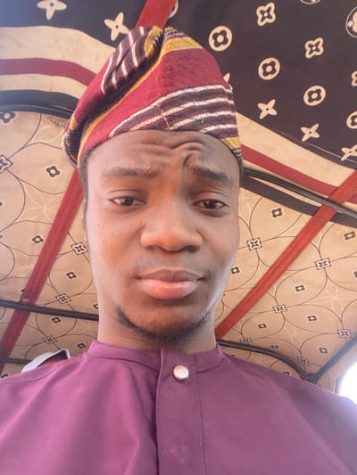 A male individual is depicted in a close-up, head-and-shoulders selfie. He has a dark complexion and wears a traditional Nigerian cap, predominantly red with yellow and white stripes. His attire includes a deep purple tunic with a mandarin collar, secured by a single, round, silver-toned button. He exhibits a slight furrow in his brow and has minimal facial hair. The subject is looking directly into the camera. The background features the interior roof lining of a vehicle, displaying a light-colored fabric with a repeating dark geometric or floral pattern. Red structural elements or straps are visible against this patterned lining. The specific city location indicated by "0801712591" in Nigeria cannot be determined from the provided numerical identifier.