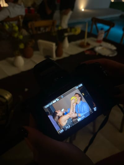 The image shows a digital camera held in a person's hand, displaying a photo on its screen. The photo on the screen depicts two people: one wearing a light blue shirt and holding a yellow object (possibly a fruit or ball) near their mouth, and another person wearing a polka-dot top. The camera settings visible on the screen include "ISO 6400" and "1/60." The camera's battery indicator shows 13339/1347.
The background of the main image (outside the camera screen) is out of focus, but suggests a dining table setting with plates, candles, decorations, and chairs. The location information, Santa Cruz Tehuixpango, Mexico, provides geographical context but is not directly visible in the image.