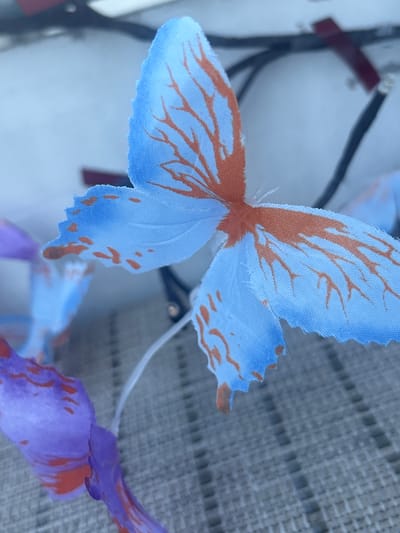 A close-up, shallow depth-of-field photograph captures a cluster of artificial butterflies, likely used as decoration or part of a costume. The prominent butterfly in the foreground has light blue wings with delicate, irregular burnt orange markings resembling veins or intricate patterns. The wing edges are frayed. Behind it, other butterflies are visible, including one with purple wings and similar orange markings, and another with light blue wings, slightly out of focus. A thin, white wire extends from the butterflies upwards, suggesting they are attached to a headband or a similar accessory. The background is blurred, showing dark wires and a red object, possibly indicating the presence of electronics or other decorative elements. The butterflies are resting on a textured surface that appears to be a grey mat or fabric, with a pattern of raised rectangular shapes. The lighting is soft, casting gentle shadows and highlighting the fabric texture of the butterfly wings. There is no indication of the specific location within Osterode am Harz, Germany.
