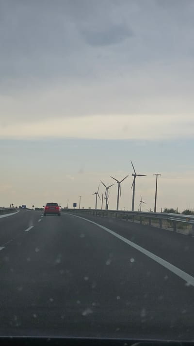 The image shows a highway with a red car in the foreground driving away from the camera. In the background, a row of wind turbines are visible on the right side of the road, suggesting a rural or industrial area. The sky is overcast, with a pale, hazy light. The highway appears to be a multi-lane road with clear lane markings. A guardrail runs along the right side of the road. The overall impression is one of travel through a landscape dominated by energy infrastructure under a cloudy sky.