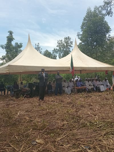 An outdoor scene in Butali Chegulo ward, Kenya, depicts a gathering under a large, off-white canopy tent with two pointed peaks. The ground consists of dry, brown vegetation and bare earth. In the background, dense green trees and foliage are visible under a light blue sky with scattered clouds.

Central to the foreground, a male speaker in a dark suit, light shirt, and dark tie stands holding a gold-colored microphone, addressing an unseen audience to his right. To the speaker's immediate right, another man stands with arms crossed, wearing a dark patterned shirt and dark trousers.

Under the tent, to the left of the speaker, several individuals are seated on blue and dark plastic chairs. One person operates a Yamaha-branded electronic keyboard. Further inside the tent, a large group of approximately 15-20 men are seated on low benches or mats, many wearing light-colored robes and white skullcaps. A green, white, and red flag, partially displaying "CHURCH," is affixed to a pole near this group. Another flag, with red and blue elements, is partially visible on the far right.

The overall scene indicates an organized outdoor event, possibly a religious or community meeting, with a speaker addressing attendees and musical equipment present. Debris, including a light-colored wrapper and a dark plastic object, is visible on the ground in the immediate foreground.