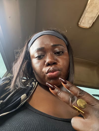 A close-up selfie of a Black woman with dark skin and long, straight dark brown hair is captured. She is wearing a dark gray headband and a black top with a ribbed texture. A black and tan patterned scarf is draped around her neck and shoulders. Her lips are pursed, and she has a nose ring. Her right hand is raised, displaying long, dark red-brown nails, and she is wearing a gold ring on her right index finger. The ring features a circular element and a horizontal bar. She appears to be in the interior of a vehicle, with a glimpse of a window and a ceiling light fixture visible in the background. The lighting suggests it is daytime. The environment is not detailed enough to definitively place it in Kugbo, Nigeria.