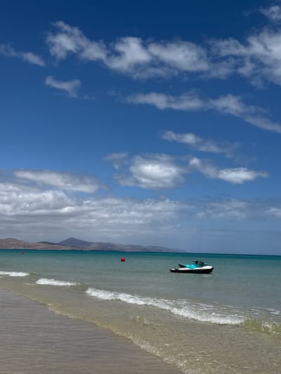 This image captures a serene coastal scene, likely in Pájara, Spain, on a clear and sunny day. The dominant feature of the upper left portion of the frame is a bright blue sky dotted with scattered, elongated white clouds, suggesting a pleasant, breezy atmosphere.
The right side of the image displays a calm, turquoise ocean meeting a light-colored sandy beach. Gentle, frothy waves are seen breaking softly along the shore, creating a diagonal line across the lower right. A white and blue/teal jet ski is floating peacefully in the shallow water, positioned near the center of the water body. Two small red spherical buoys are visible in the water; one is closer to the jet ski, and another is further down the beach towards the foreground.
In the background, a range of mountains or hills rises, their arid, light brown slopes indicating a dry climate. The mountains appear somewhat hazy in the distance. The overall impression is one of tranquility and leisure, with no human activity directly depicted. The time of day appears to be mid-day given the bright lighting. No visible text is present in the image.
