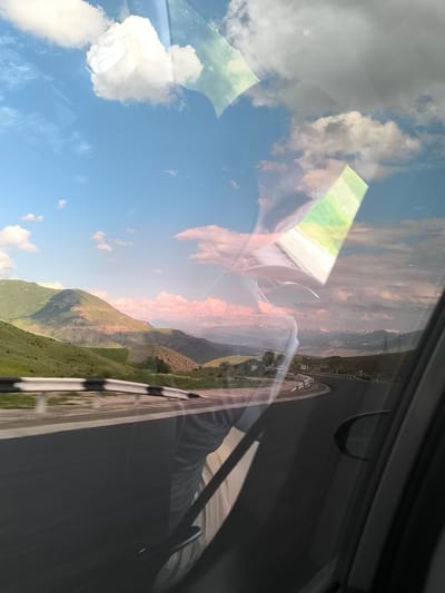 The image captures a scenic mountainous landscape from the perspective of a moving vehicle, looking out a side window. The setting is Yelpin, Armenia.
In the foreground, a paved road curves to the right, bordered by a black and white striped guardrail. The road appears slightly blurred, indicating the car's movement. Lush green hills and mountains dominate the midground and background, with some slopes showing visible rocky textures and varying shades of green vegetation. Portions of the mountains are illuminated by warm sunlight, while others are cast in shadow. In the far distance, snow-capped mountains are faintly visible under a more overcast sky on the right side of the frame.
The sky overhead is partly cloudy, featuring patches of clear blue interspersed with white and light pink clouds, suggesting either early morning or late afternoon/early evening light.
A significant feature of the photograph is the heavy reflections and refractions on the car window. These include distorted shapes of the car's interior, possibly the dashboard or a passenger, and bright, geometric light flares or reflections on the upper and central parts of the window. The car's door frame and side mirror are partially visible on the far right edge of the image. The overall effect is a layered view, combining the external landscape with internal reflections, conveying a sense of a road trip or journey through rugged, beautiful terrain. No discernible text is visible.