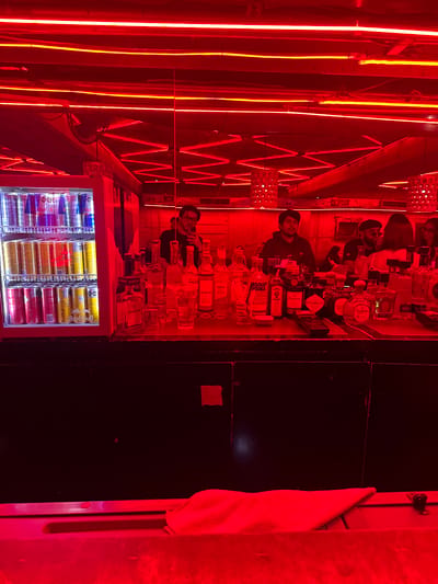 The image shows a bar interior, likely in Guadalajara, Mexico, judging by the brands of alcohol and Red Bull flavors stocked in the refrigerator. The lighting is predominantly red with yellow neon accents. A bar counter displays multiple alcohol bottles including brands such as Absolut Vodka, Hendrick's Gin, Don Julio Tequila, and a few others like Desaronno and 45 Conejos. A bartender stands behind the counter, possibly serving or preparing drinks for patrons. The foreground features a refrigerator filled with Red Bull energy drinks in different flavors. A black panel wall is seen towards the right, and a white towel hangs nearby. Patrons and other staff members are visible behind the bartender.
