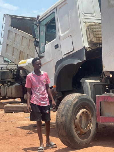 A young man stands near the front of a white dump truck in Kwali, Nigeria. The truck's cab is partially visible, as is one of its large front tires. The man wears a pink t-shirt and black shorts and gray sandals. The ground is brown dirt. The sky is blue.