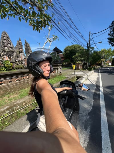 A young woman wearing a helmet is riding a scooter in Petulu, Indonesia. She is positioned on the right side of the road, next to a raised concrete curb with grass on the other side of the curb. There is a dark gray asphalt road with a solid white line marking the edge. In the background on the left is a traditional Balinese temple structure. A tangle of utility wires stretches overhead against a blue sky.