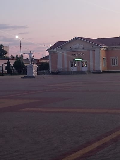 The image captures an outdoor scene at dusk or dawn in Panino, Russia, with a prominent statue of Vladimir Lenin in the foreground. The statue, made of a silver-colored material, depicts Lenin with his right arm extended, pointing forward. It stands on a pedestal. Behind the statue, a building with "ВОСТОК" (Vostok) written in Cyrillic letters is visible. The building is painted in pastel colors with white vertical stripes and has a dark roof. It appears to be a public building, possibly a community center or a railway station, given the context of the name "Vostok" which can mean "East." Several streetlights are present in the scene, with one illuminating the sky. The ground in the foreground is paved with interlocking bricks, featuring yellow and red striped patterns. The sky displays a soft gradient of pink and purple hues, indicating twilight. Other structures and trees are visible in the background, suggesting an urban or semi-urban environment. There are no other individuals or animals clearly discernible in the image.