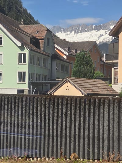 This is a vertical, landscape-oriented photograph taken from an elevated viewpoint looking down and across a village. The scene is dominated by a range of snow-capped mountains in the background, under a clear blue sky with a few wispy clouds. In the foreground, the roofs of several buildings are visible, featuring a mix of brown tile and gray corrugated materials. One prominent building in the mid-ground is painted a pale green and has numerous white-framed windows. Another building is a warm orange hue. Dense green trees are scattered throughout the village. To the right of the frame, a dark, textured surface with horizontal lines, possibly a corrugated metal fence or wall, occupies a significant portion of the image. The overall impression is a picturesque, sunny day in a mountainous European village, likely in Switzerland, given the location context of Göschenen. There is no discernible text within the image.