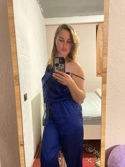 A woman is taking a mirror selfie in a dimly lit room. She is wearing a blue satin jumpsuit with black lace trim. Her hair is blonde and wavy. She is wearing rings on her fingers. The room has white brick walls and a textured wall. There is a bed with a light blue comforter in the background. A rug with a red and black pattern is on the floor. The overall scene appears to be in a residential setting.