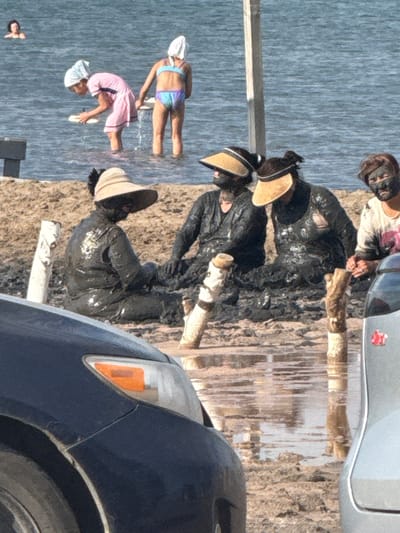 The image captures a scene on a muddy shoreline, likely near the city of Yamyishevskoye rural district, Kazakhstan. In the background, the vast expanse of a body of water is visible. In the mid-ground, two young girls are standing in the water. One, wearing a pink swimsuit and a white headscarf, is bending down, holding a white object. The other, in a colorful swimsuit and white headscarf, stands beside her. Further forward, three individuals are seated on the muddy ground. They are covered in dark mud, and two of them are wearing wide-brimmed sun hats. The person on the far right is wearing a t-shirt with a floral pattern and also has mud on their face and neck. The surrounding environment is characterized by mud, with several wooden posts sticking out of the ground, some submerged in shallow water. In the foreground, parts of two vehicles are visible; a dark-colored car on the left and a lighter-colored vehicle on the right. The overall impression is of people engaged in a mud bath or similar activity by the water.
