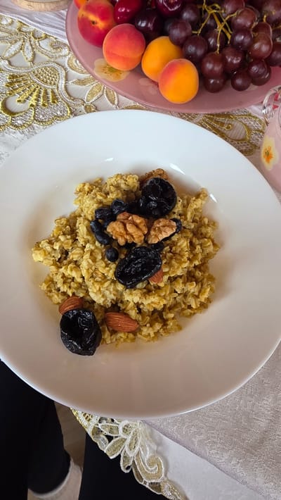 The image shows a white plate filled with oatmeal topped with walnuts, prunes, and blueberries. In the background, a pink plate of fruit, including apricots and grapes, is visible on a table with a decorative cloth. The scene appears to be an indoor setting, possibly a home or a cafe, in the village of Kupoyarovo, Russia.