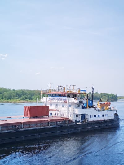 The image shows a large barge carrying a shipping container on a waterway. The barge is white with a black hull and has a multi-level superstructure. The red shipping container is secured on the deck. The waterway is blue with subtle ripples. Lush green trees line the banks in the distance. The sky is a light blue with sparse clouds. Based on the identified location (Нифантово, Russia), this scene is likely the Kama River or a similar waterway used for transport in the area.
